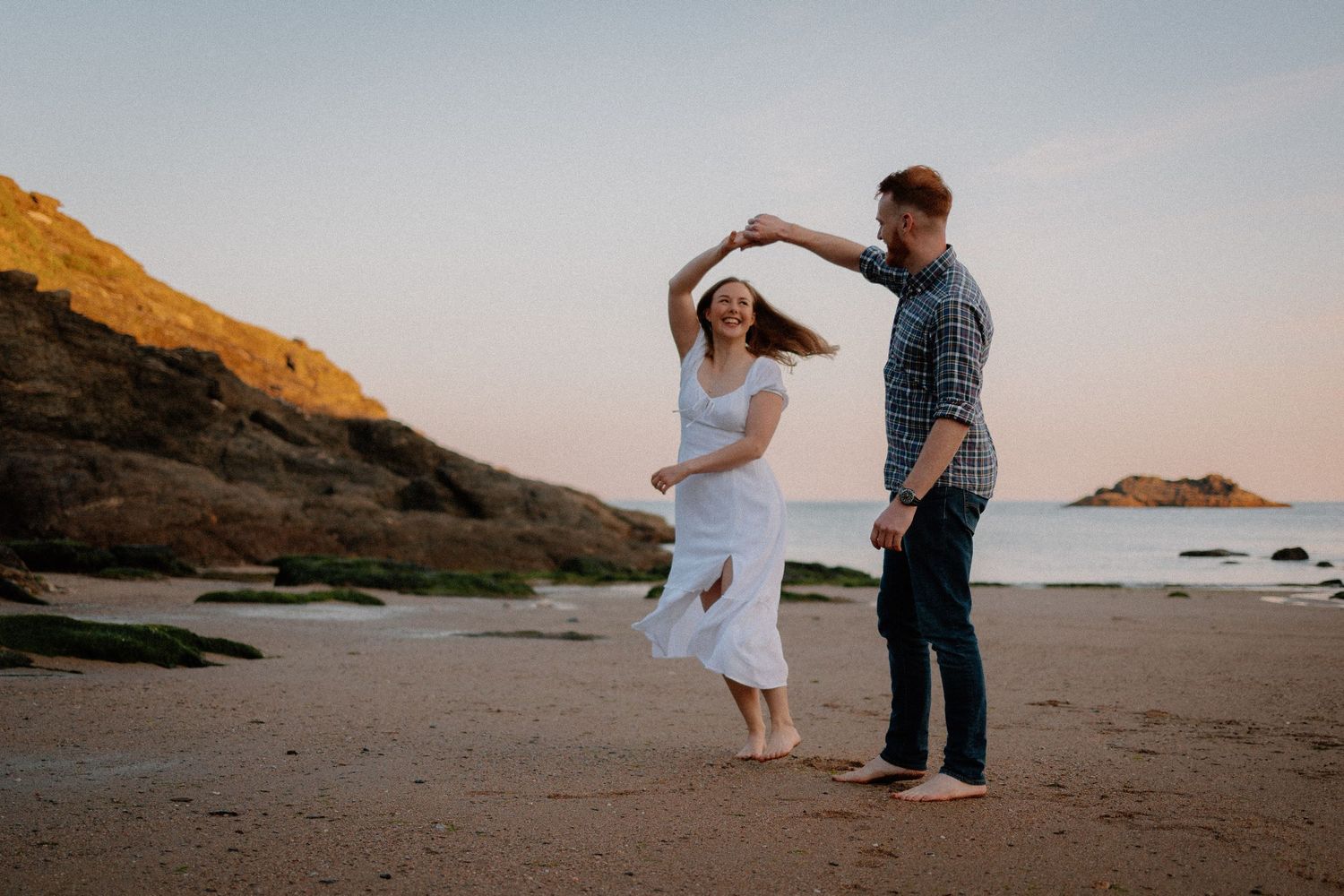 A couple dances together on a scenic beach at sunset with mountains in the background.