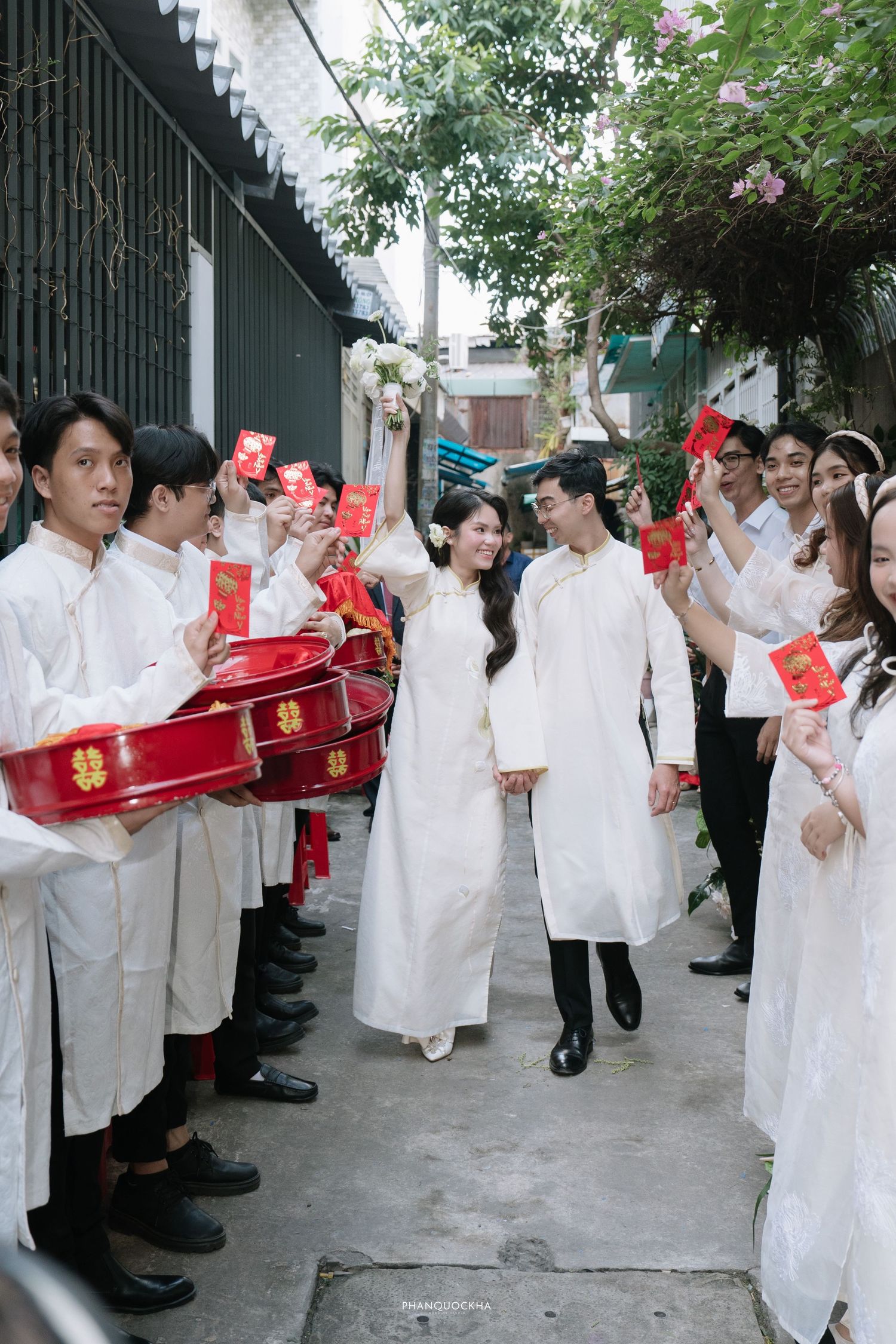 Bride and groom in white ao dai walk through a ceremonial procession with guests holding red envelopes.