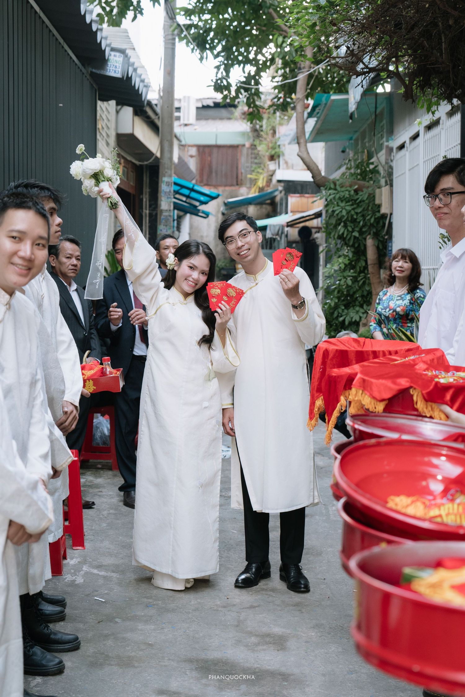 Vietnamese wedding couple in white ao dai holding red envelopes celebrating outdoors with guests.