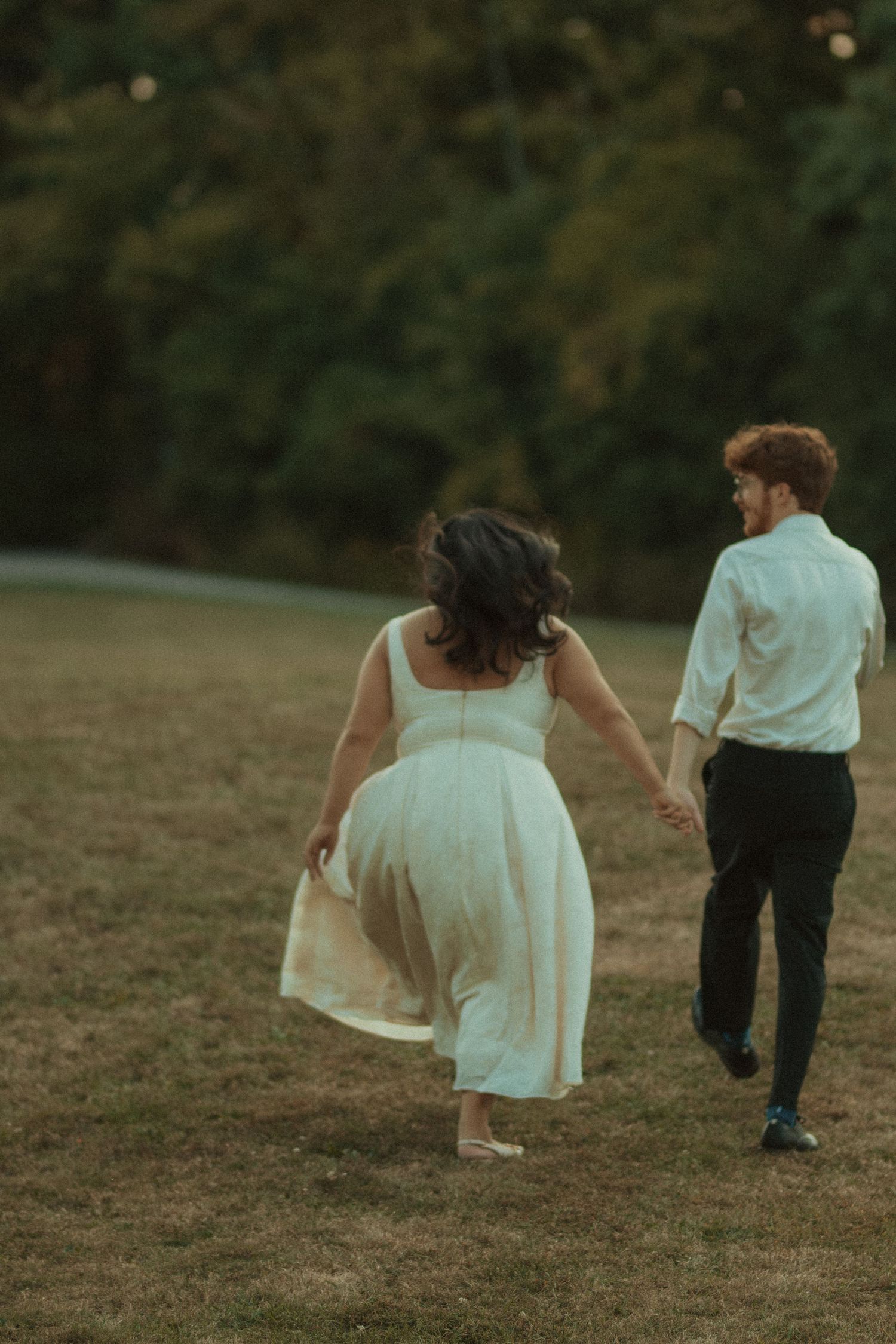 A couple holds hands while running through a lush green field, the woman in a white dress and man in dress shirt and trousers.