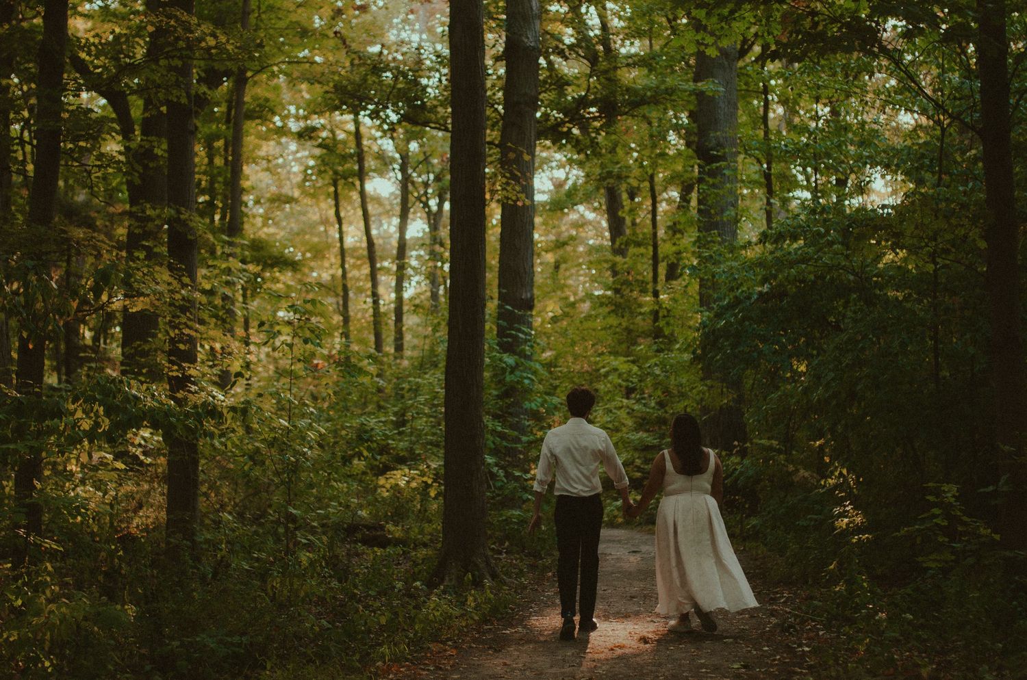 A couple in wedding attire walks hand-in-hand down a forest path surrounded by tall trees and lush greenery.