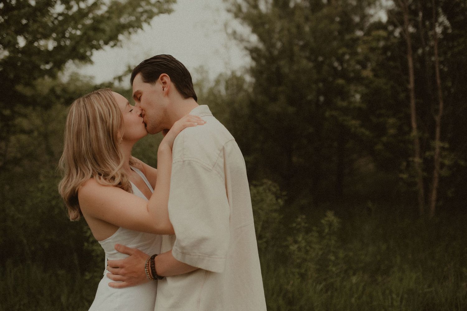 A couple shares a romantic kiss outdoors surrounded by lush green trees in a soft, dreamy engagement photo.