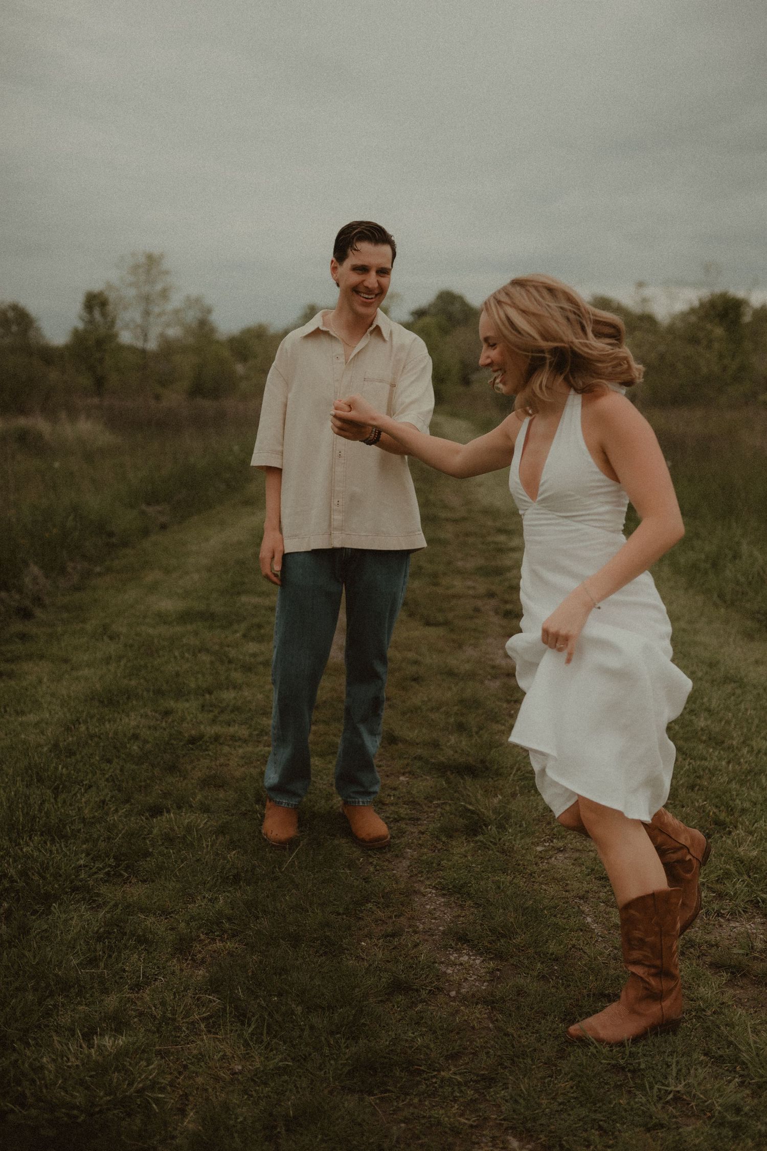 A couple dances in a green field, woman in white dress and cowboy boots, man in beige shirt and jeans.
