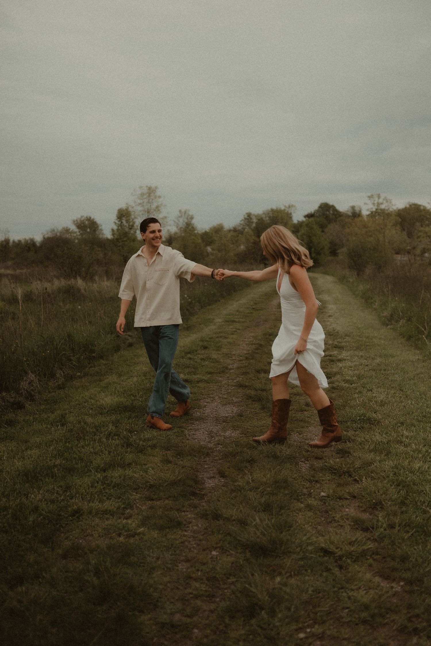 A couple holds hands and dances on a grassy path in a field, wearing casual country attire including cowboy boots.