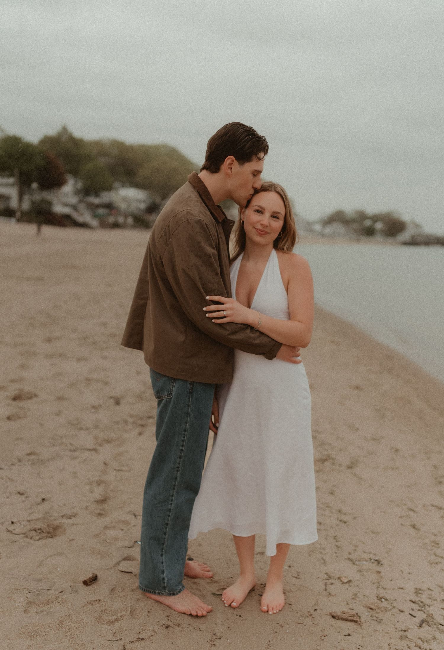 Couple embracing on sandy beach, man kissing woman's head, she wears white dress, cloudy sky backdrop.