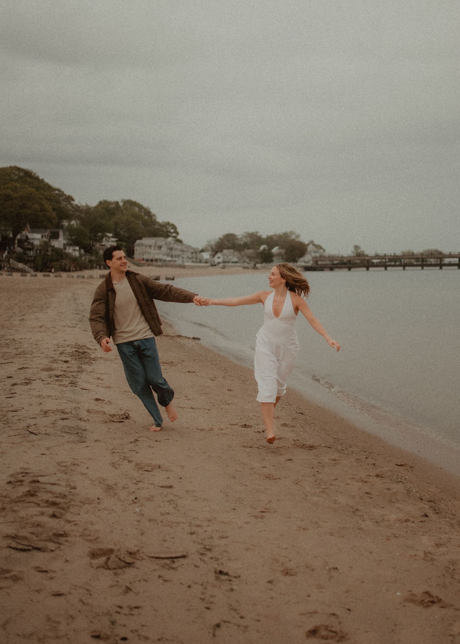 Couple holding hands while walking on beach at sunset, man in casual wear and woman in white dress near waterfront.