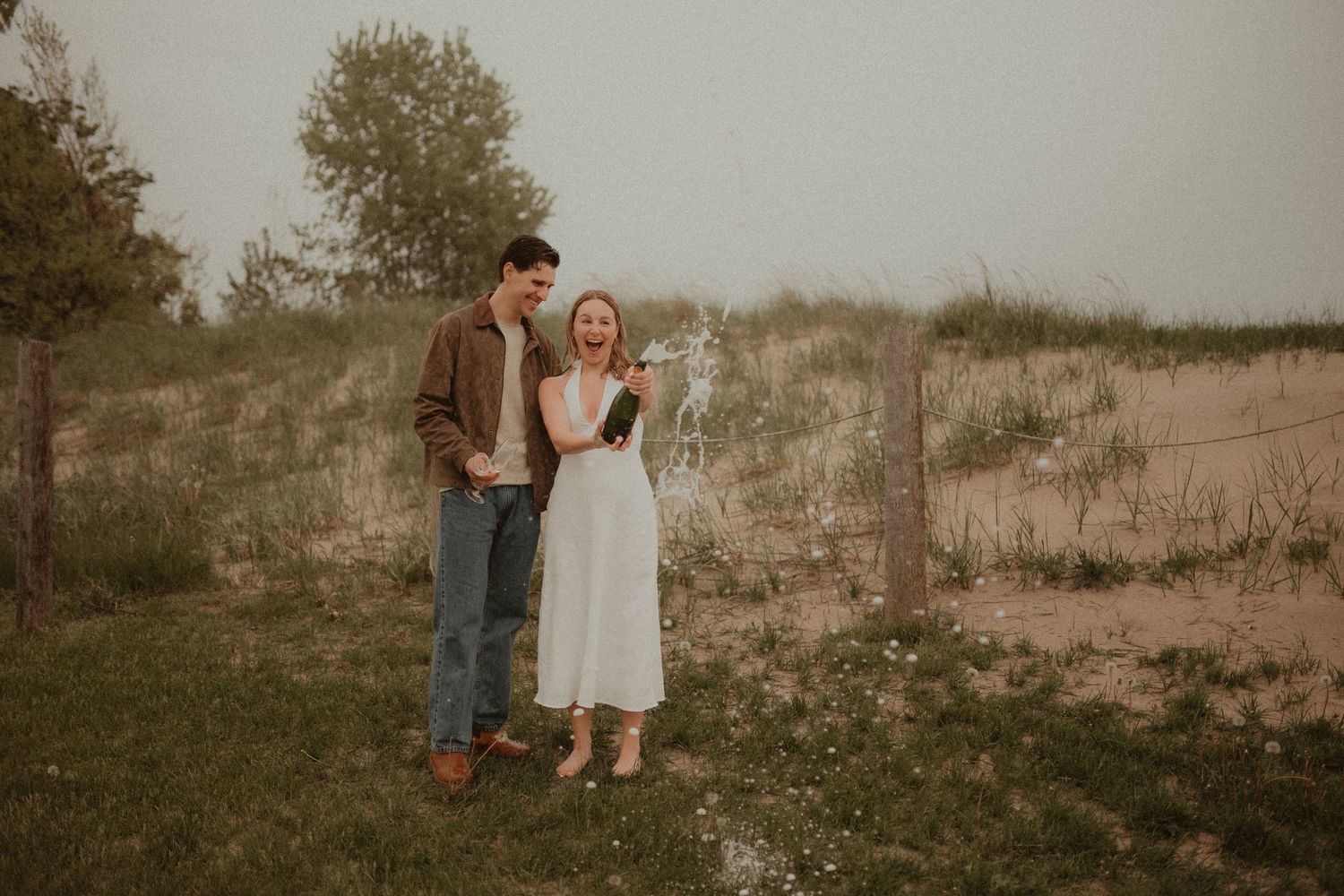 Couple smiling together outdoors near sand dunes, woman in white dress holding wildflowers, man in brown jacket and jeans.