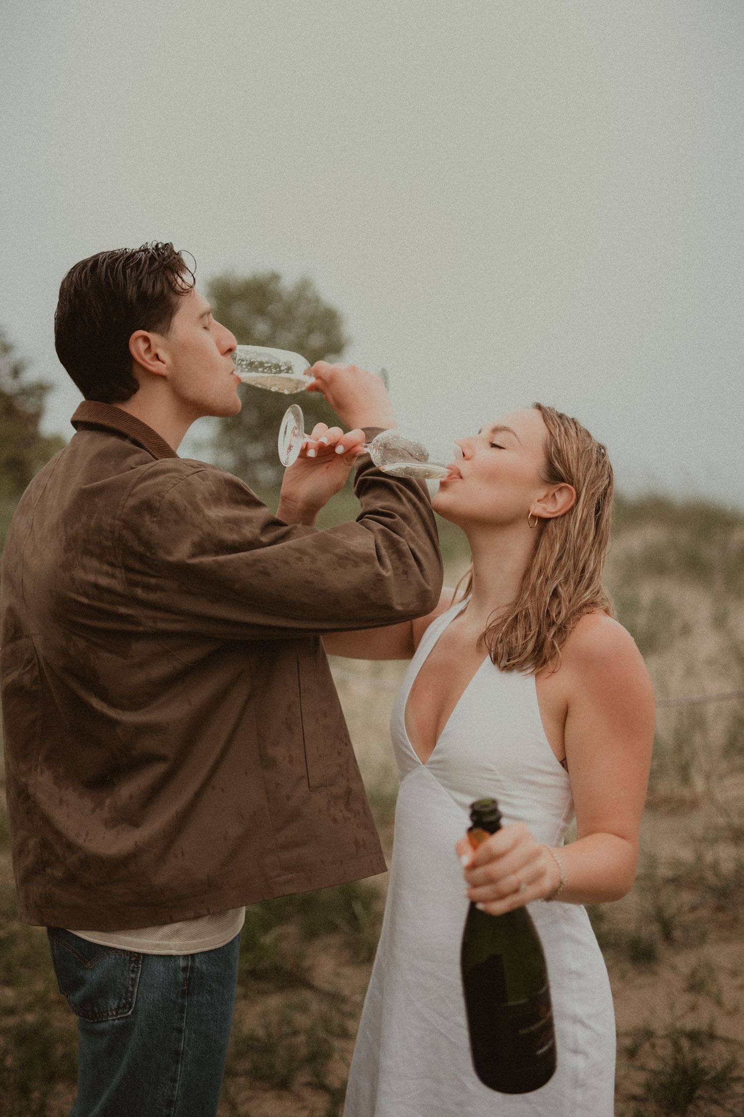 Couple drinking champagne from glasses outdoors in sandy dunes, woman in white dress holding bottle, man in brown jacket.