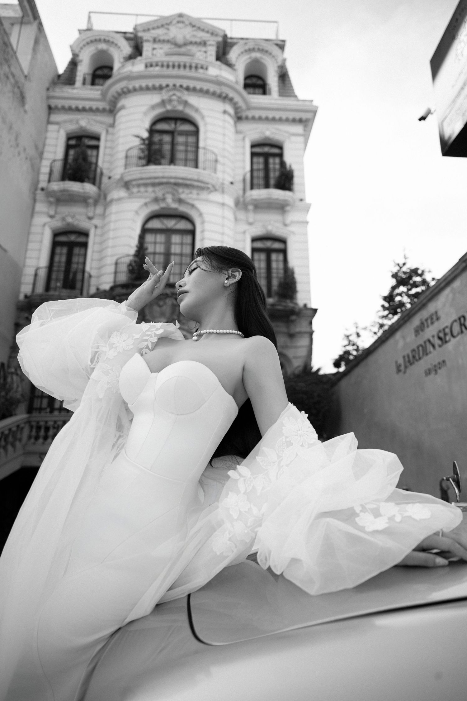 Black and white photo of bride in voluminous white gown posing on vintage car outside historic building.