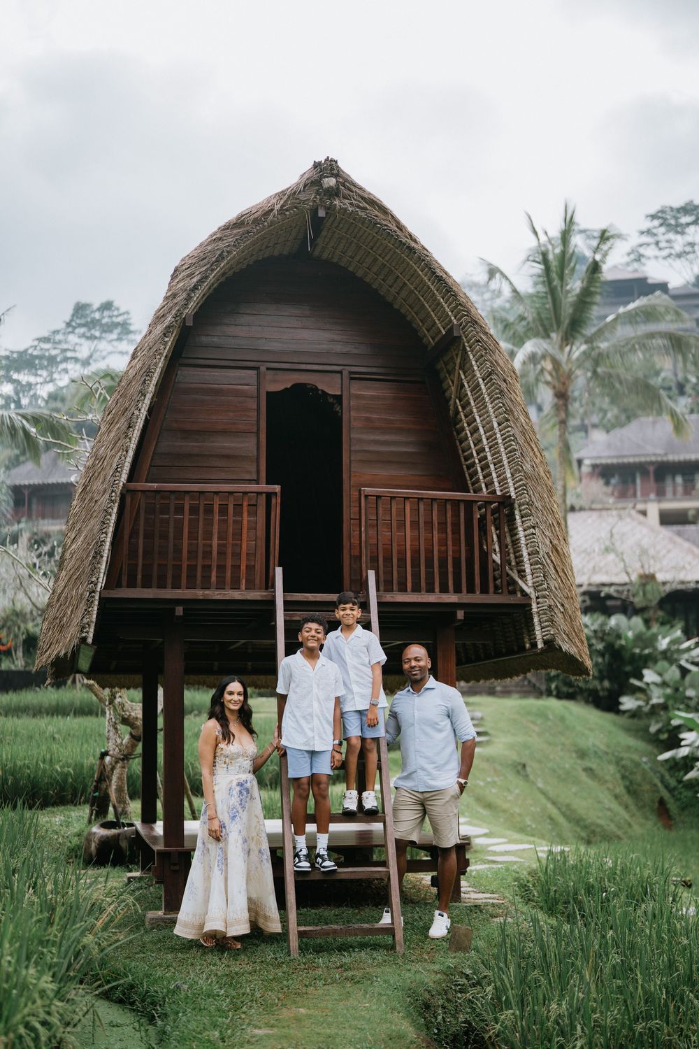 Family poses in front of traditional thatched-roof Balinese huts surrounded by rice fields.