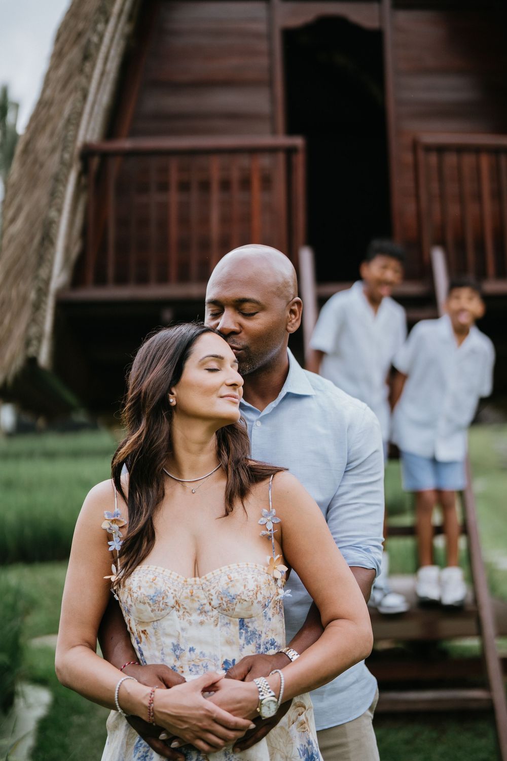 A romantic couple embraces in front of a rustic wooden cabin during a tropical photoshoot.