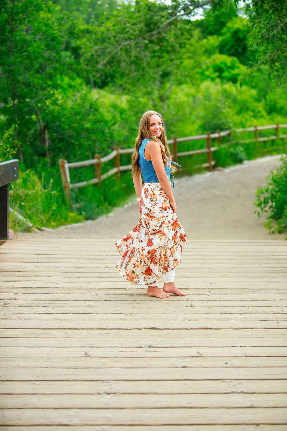 A person in a floral summer dress twirls playfully on a wooden boardwalk surrounded by lush green foliage.