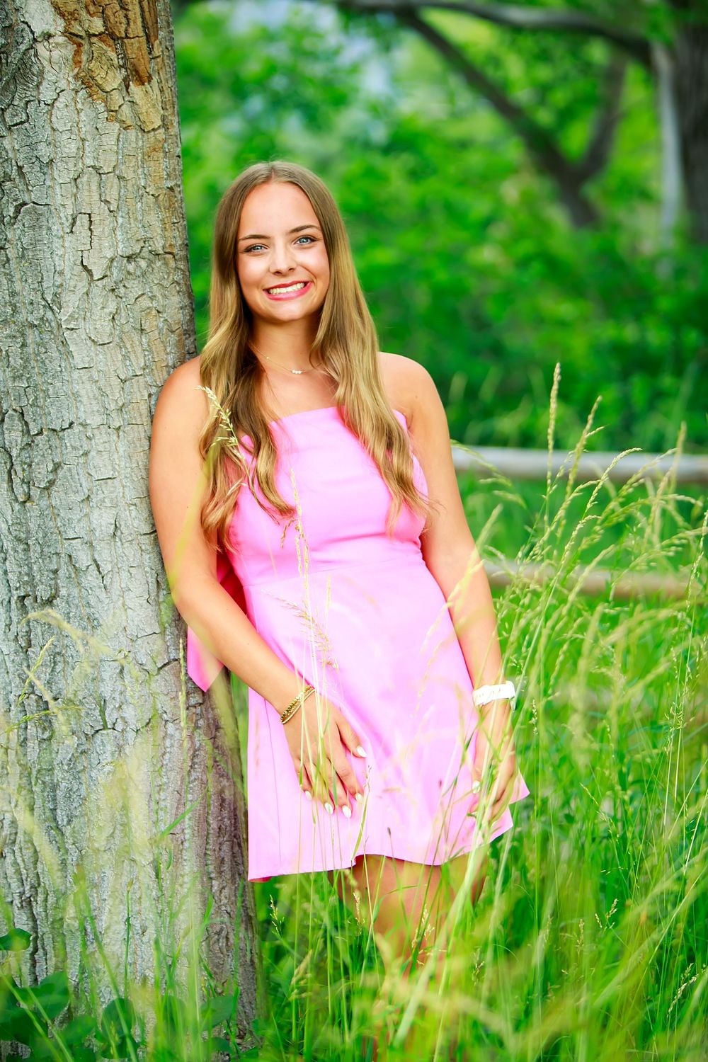 Person in a pink sleeveless top poses in tall grass near a tree trunk in a vibrant summer setting.