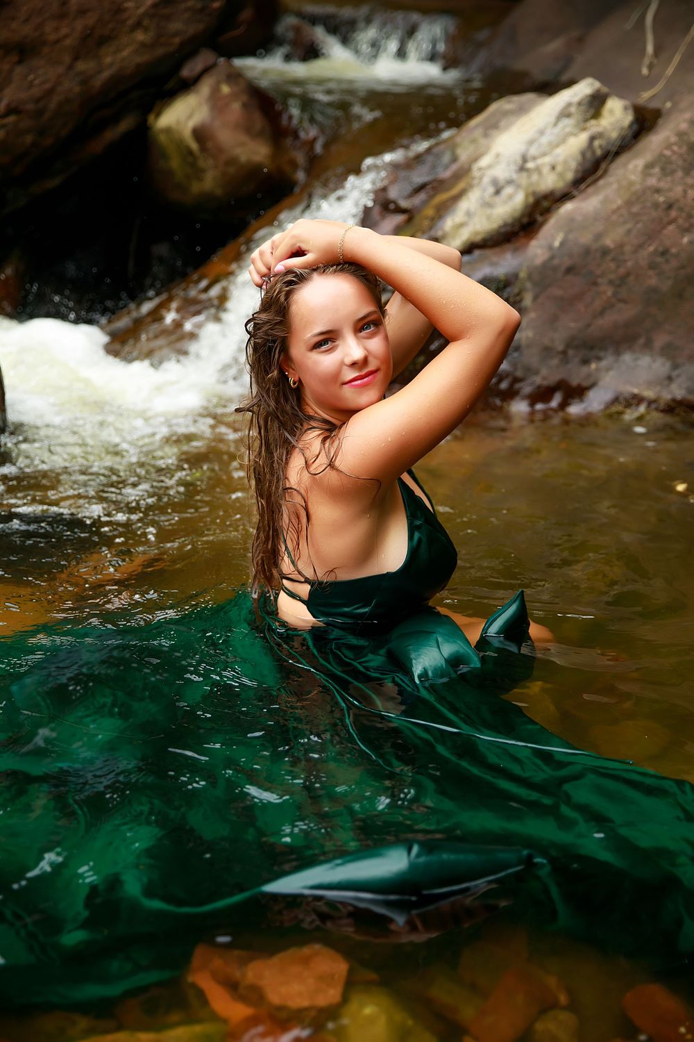 Figure poses in emerald green dress while floating in natural waters beside rocky formations.
