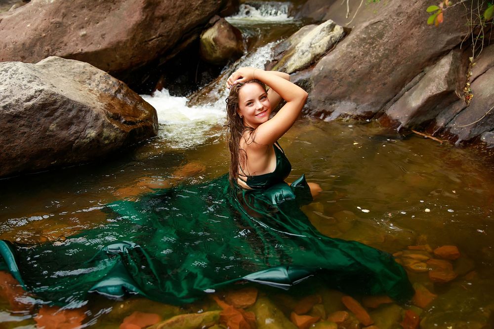 A flowing emerald green dress billows in clear water against rocky formations near a waterfall.