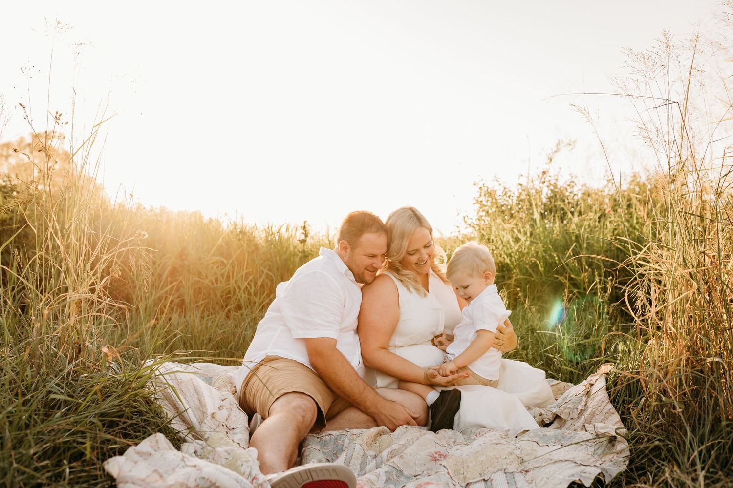 Family cuddling together on white blanket in golden field at sunset.