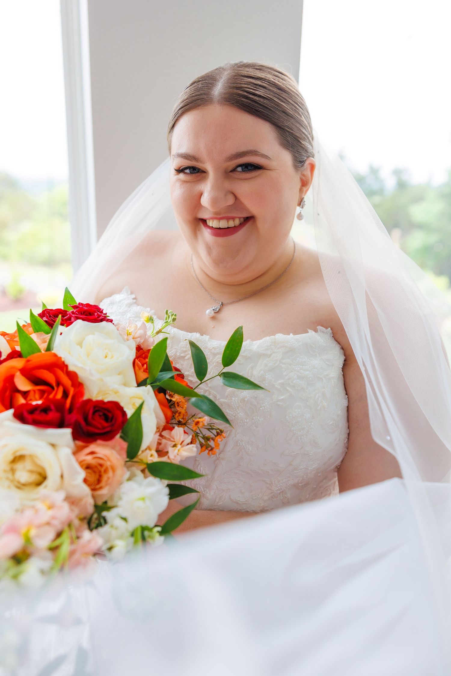 A wedding bouquet featuring vibrant red and orange roses with green foliage against a white bridal gown at the Atrium in Branson, MO.