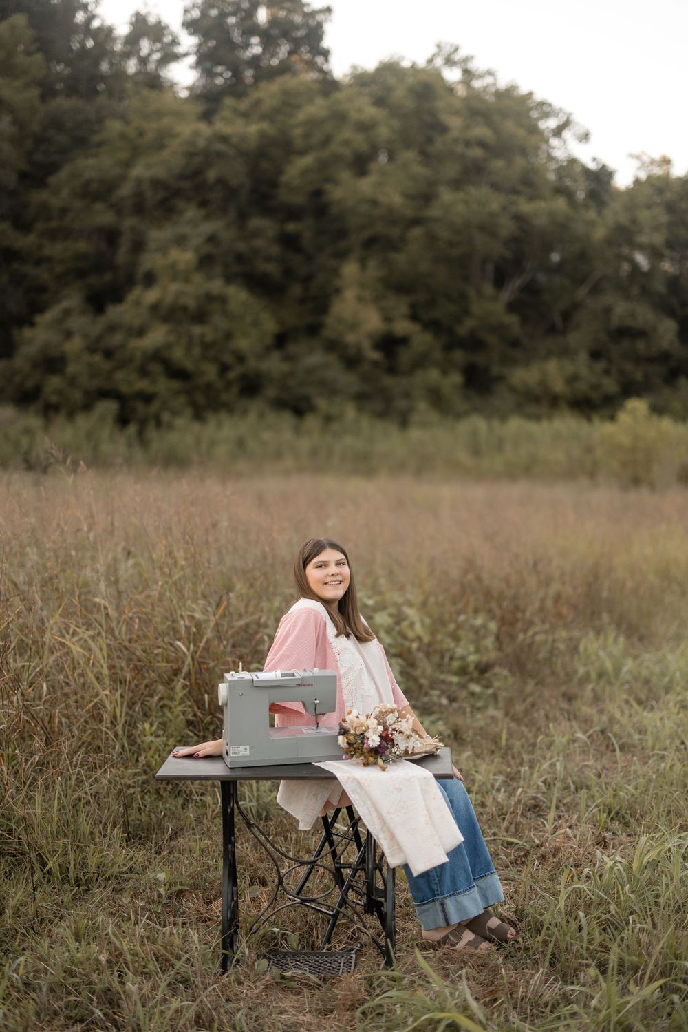 Rustic outdoor scene with typewriter and flowers on desk in tall grass field with trees in background.