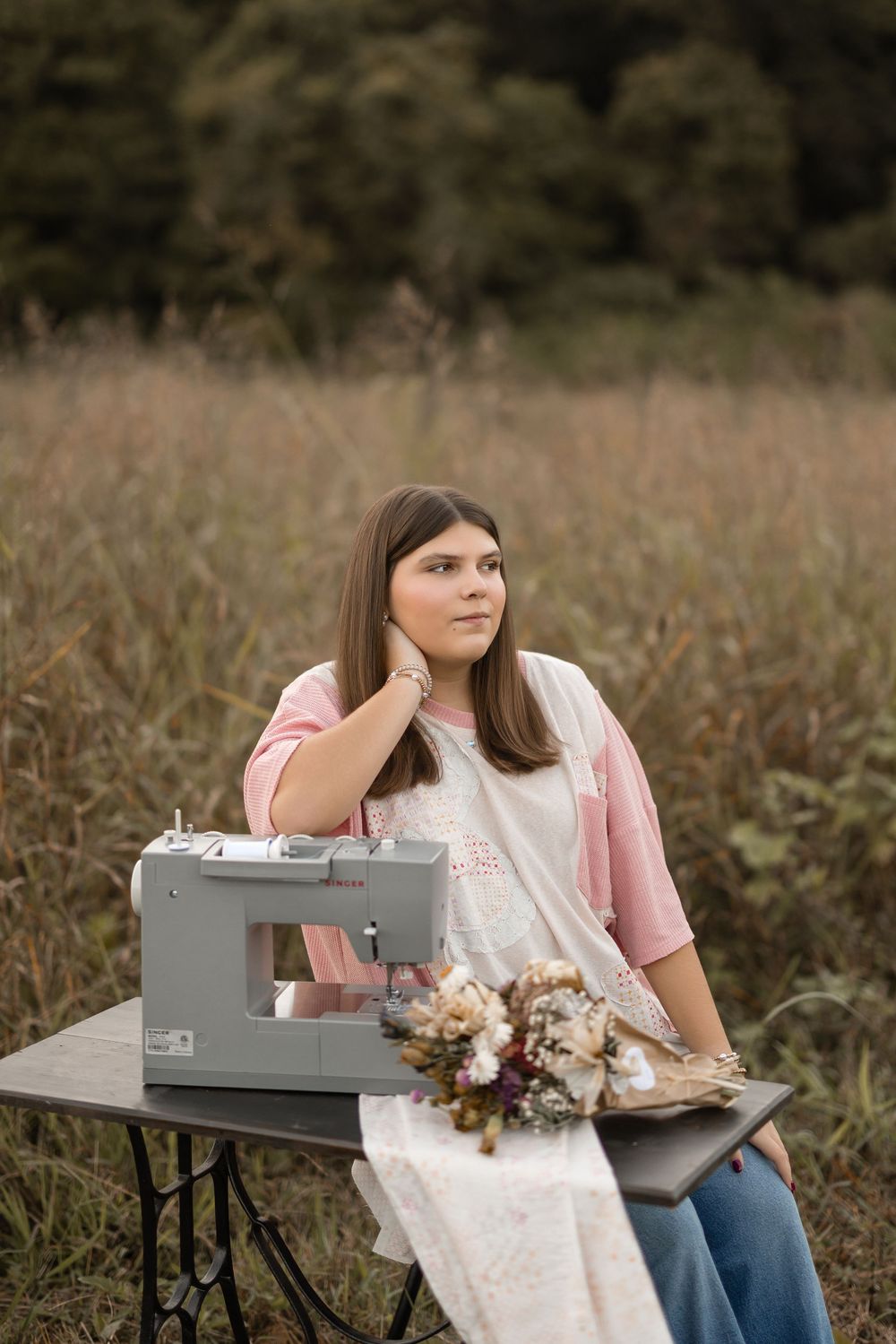A person in pink and white sits at a rustic outdoor desk with dried flowers and a vintage typewriter.