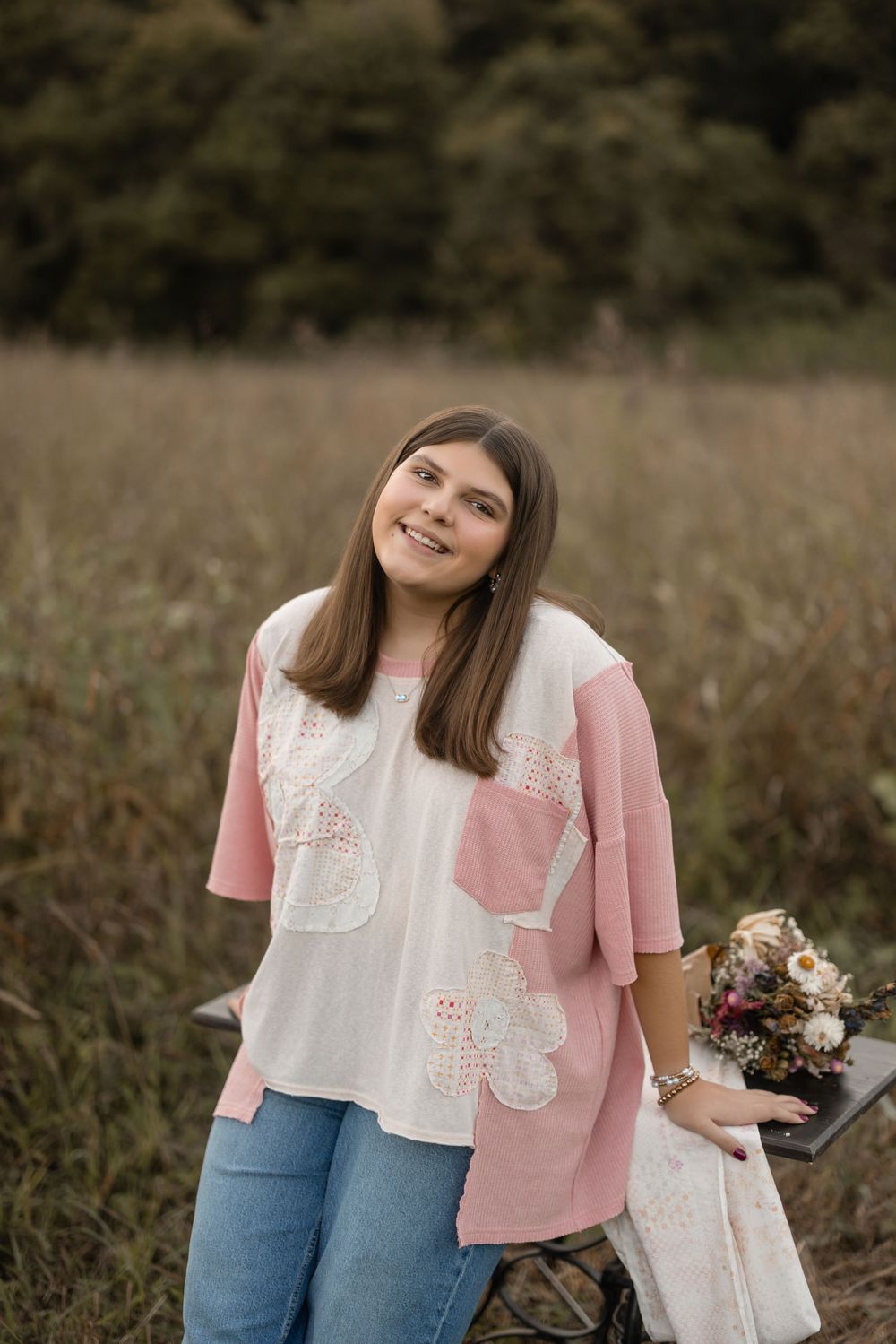 A person wearing a pink shirt and blue jeans holds flowers while standing in a field during an outdoor photo session.