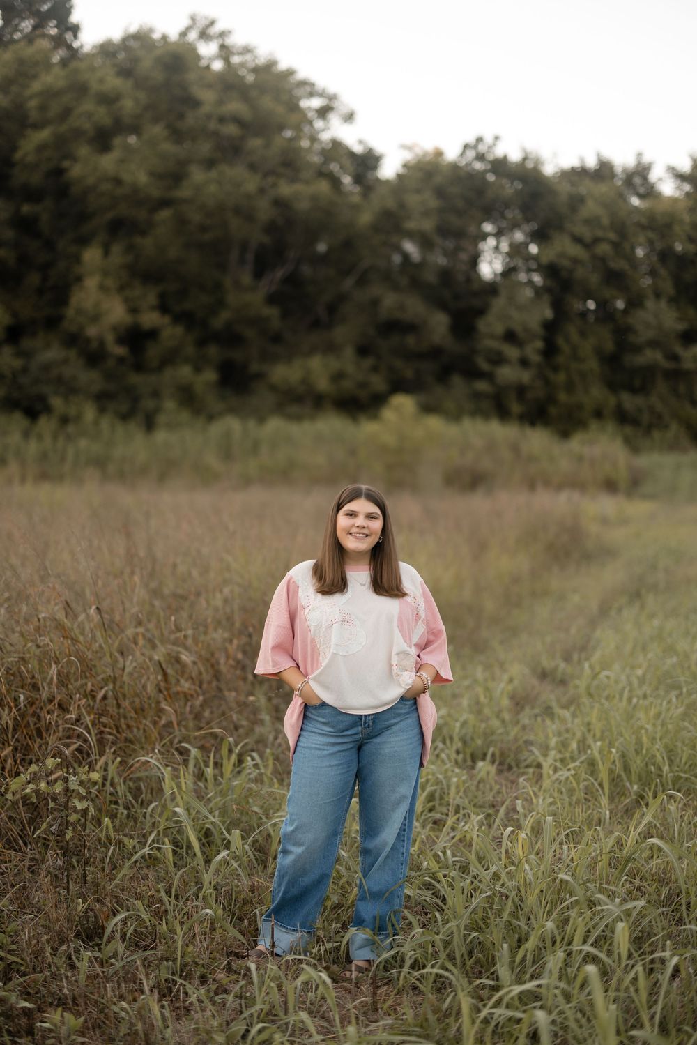 Someone poses in a grassy field wearing a pink top and blue jeans on a sunny day.