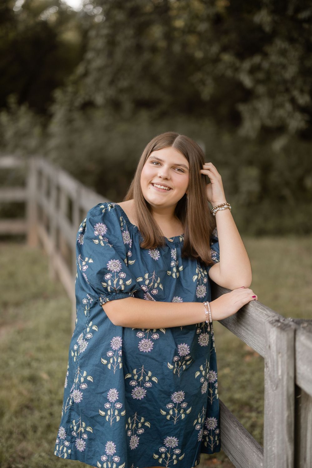 Woman in blue floral dress leans on white wooden fence post during outdoor portrait session on sunny day.