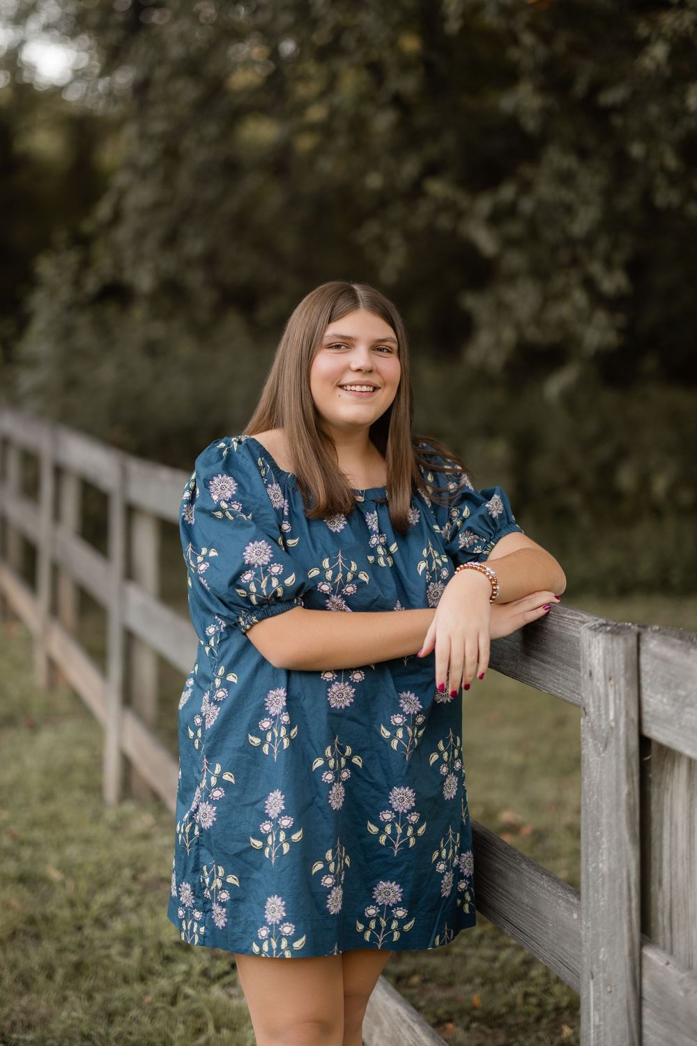 Woman in a blue floral dress poses against a wooden fence in an outdoor setting during golden hour.