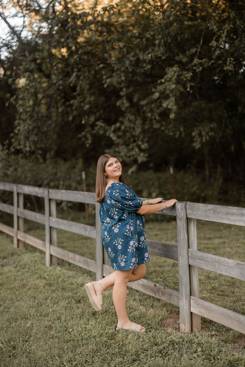 A woman in a blue floral dress stretches her leg on a wooden fence in an outdoor country setting at sunset.