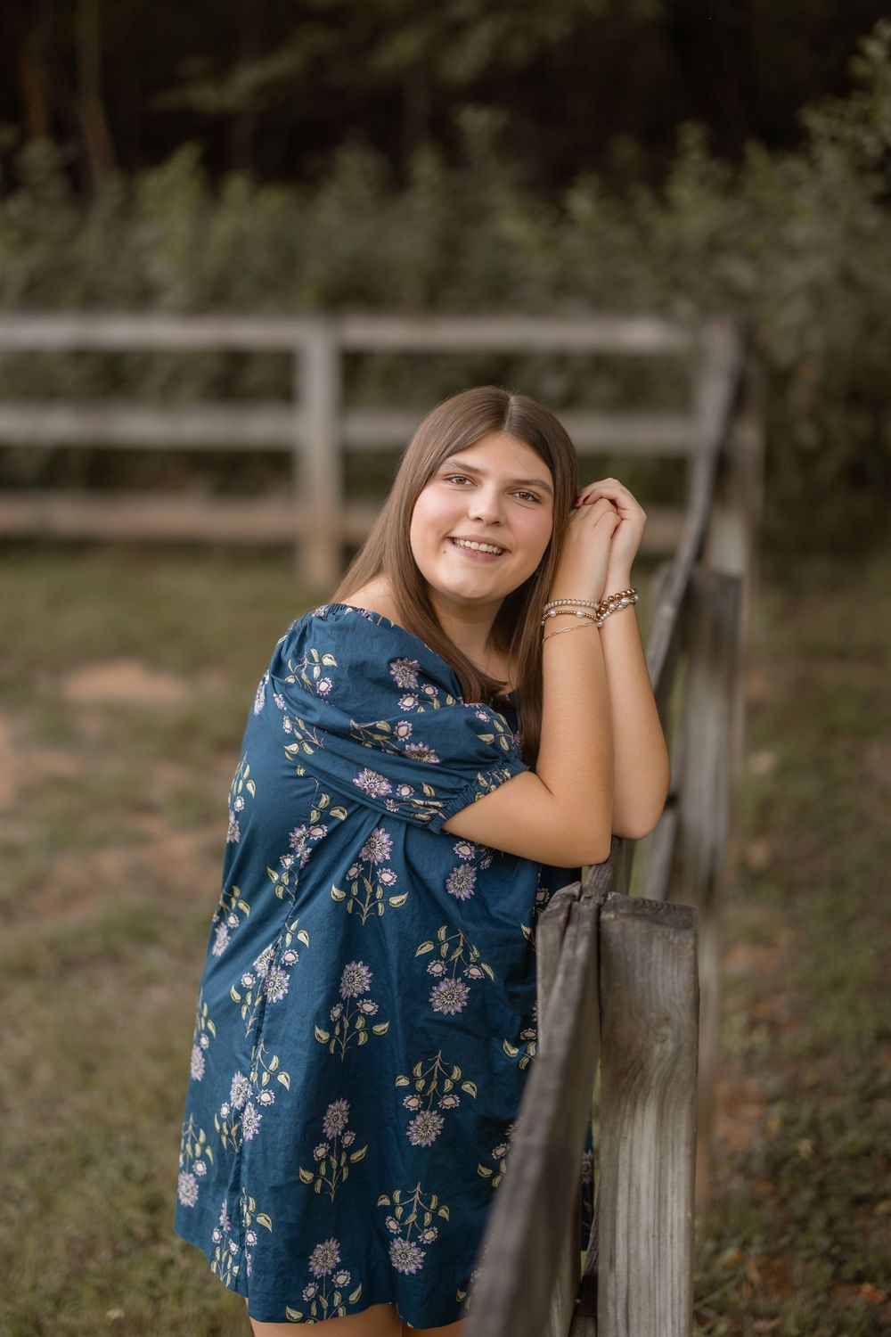 Person in blue floral dress poses outdoors against wooden fence in natural rural setting.