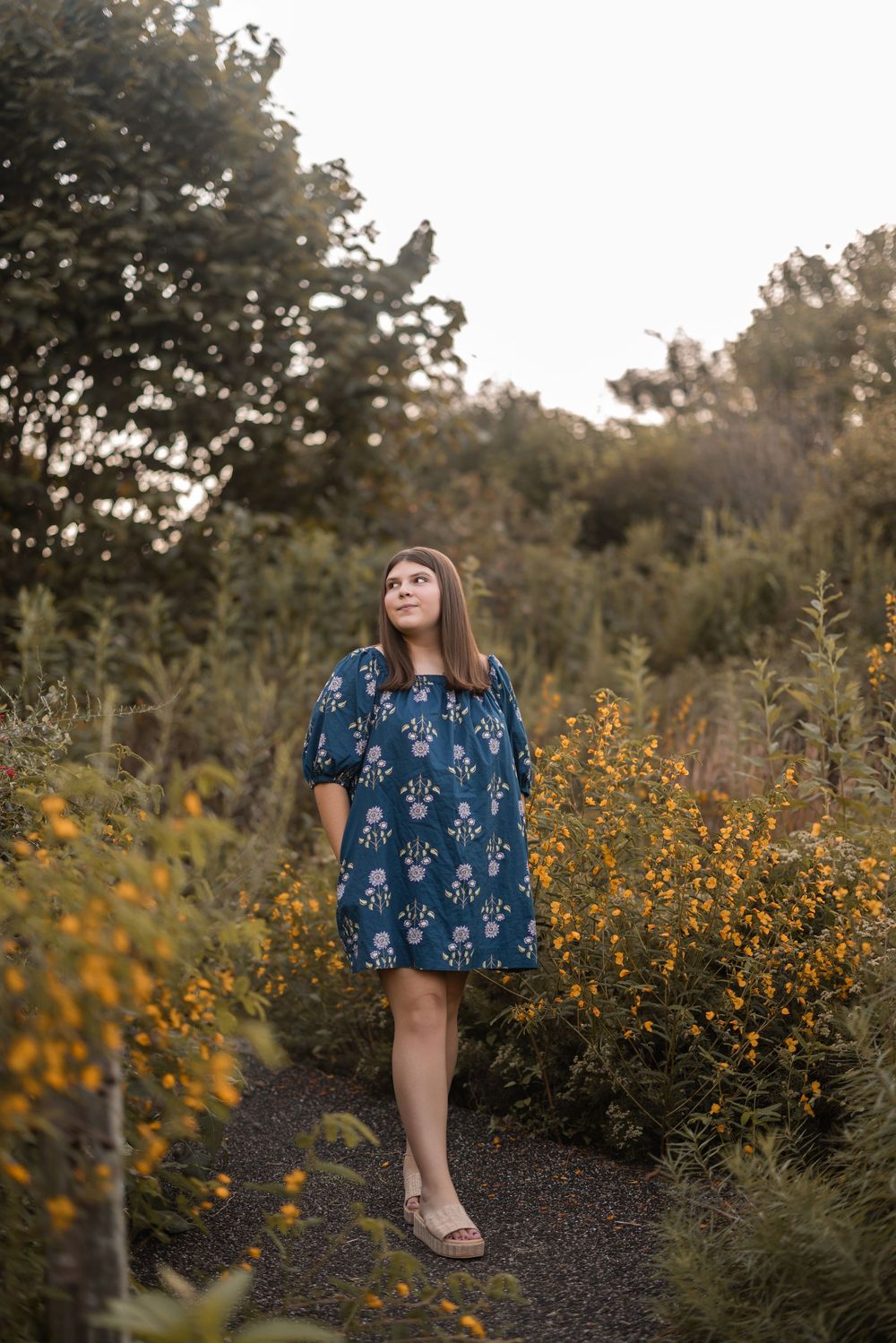 Yellow wildflowers bloom in a garden while a person in a blue floral dress poses among the natural scenery.