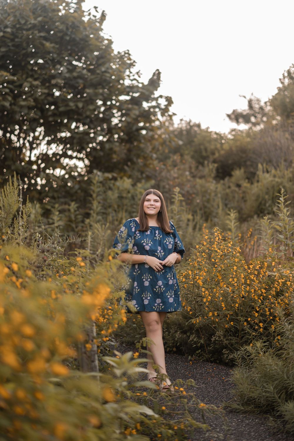 Person in blue floral dress poses near yellow wildflowers at golden hour in outdoor garden setting.