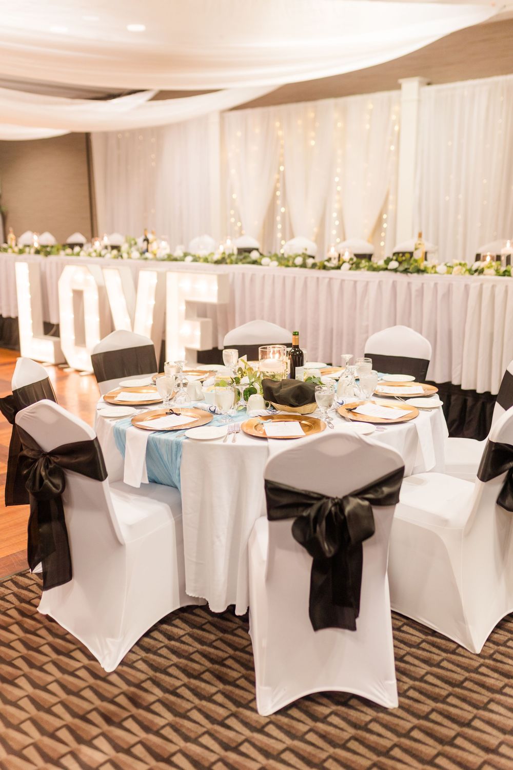 Elegant wedding reception table setup with white spandex chairs, black sashes, and LOVE letters at head table.