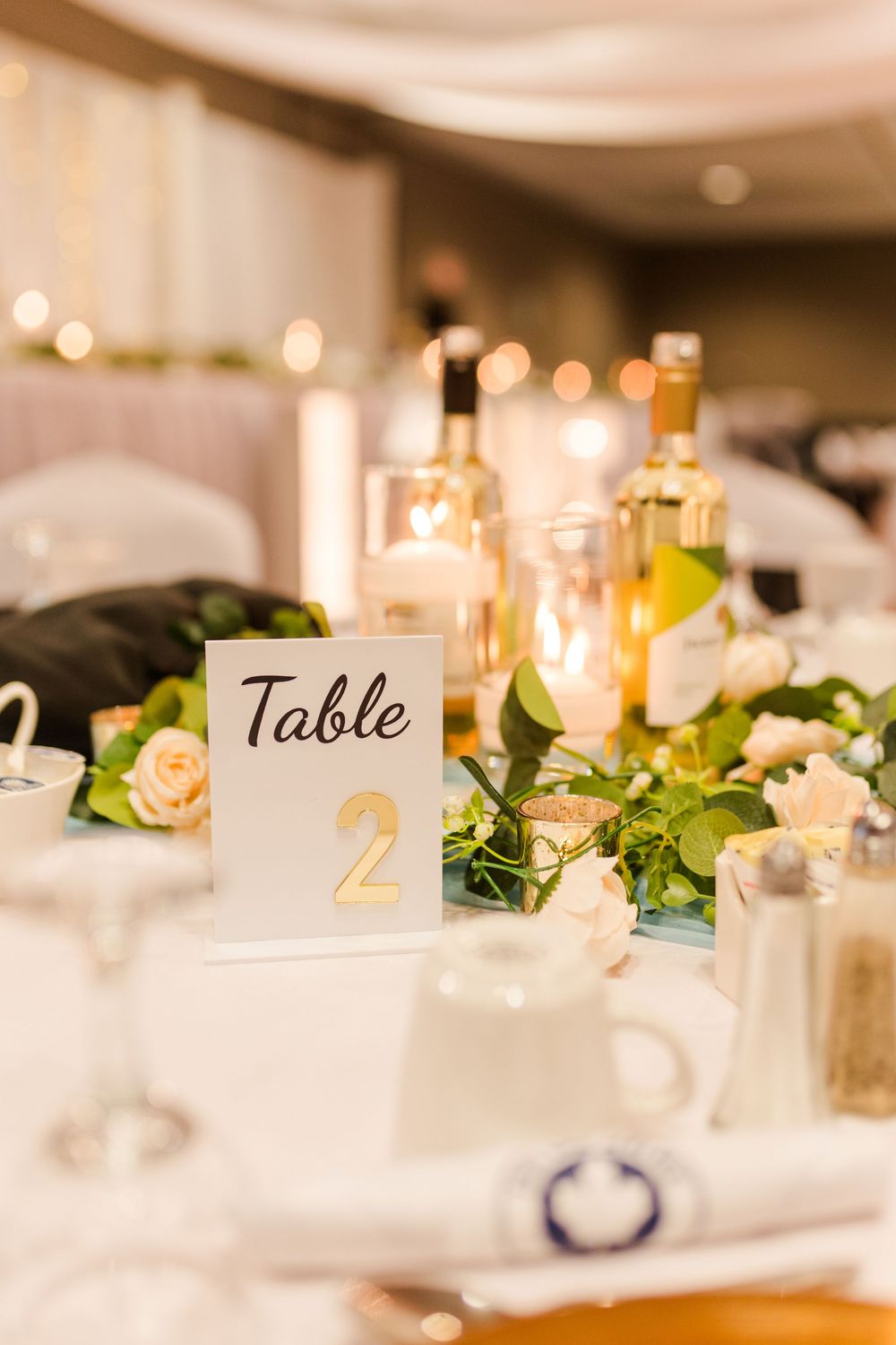 Elegant wedding reception table setting with gold table numbers, candles, and peach roses against a blurred background.