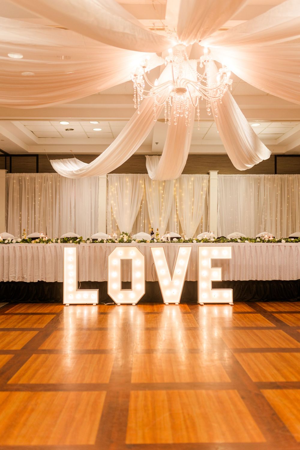 An elegant wedding reception hall with white draped ceiling and illuminated LOVE letters on a polished wood dance floor.