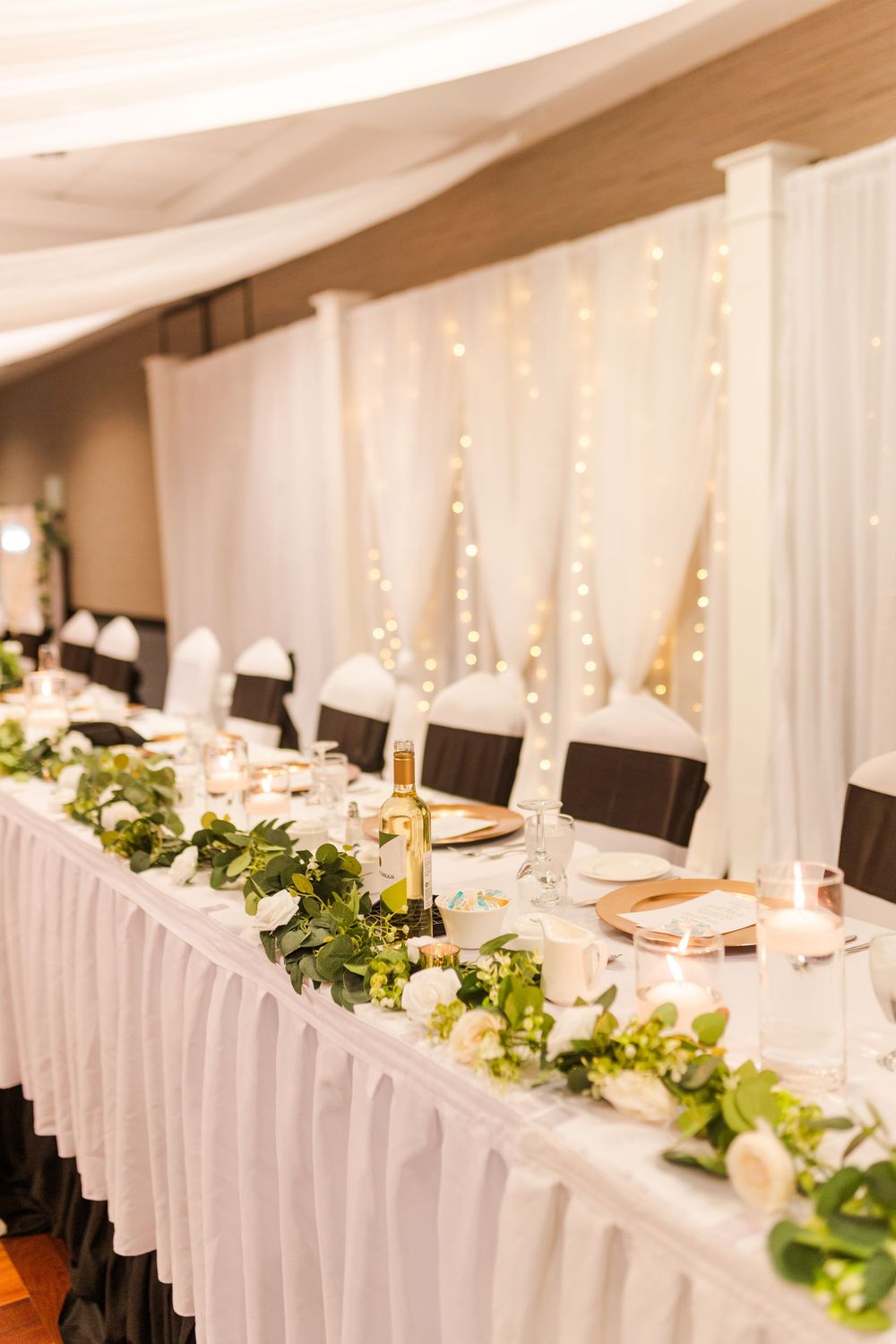 Elegant wedding reception table setup with white linens, twinkling lights, and fresh greenery garlands.