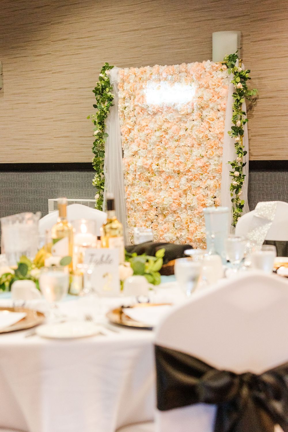 Wedding reception dining table setup with floral wall backdrop and white linens in elegant event space.