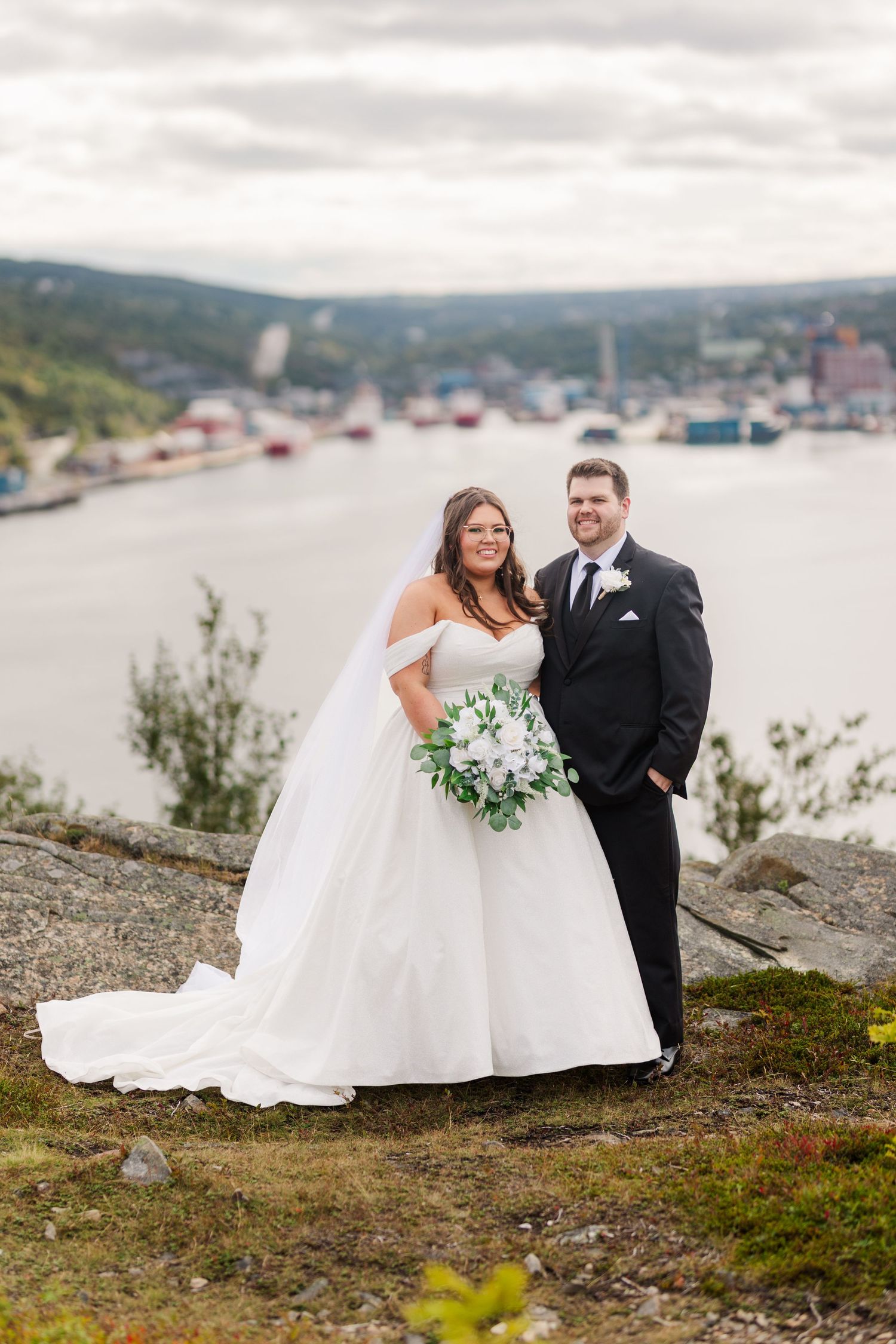 A newlywed couple shares tender moments on a scenic hillside overlooking a waterfront harbor landscape.