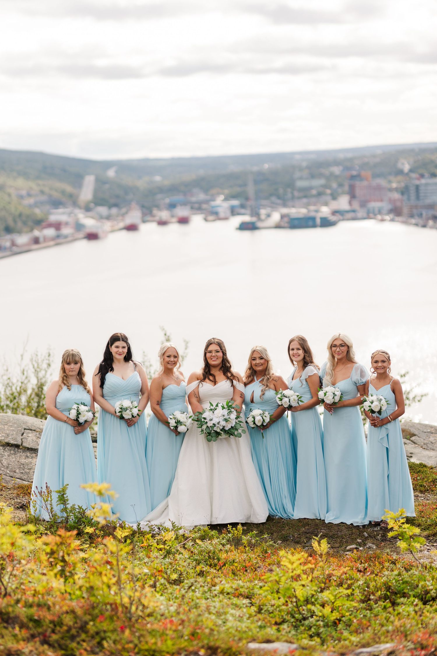 Wedding party wearing light blue bridesmaid dresses pose together on a scenic coastal hill with ocean views.