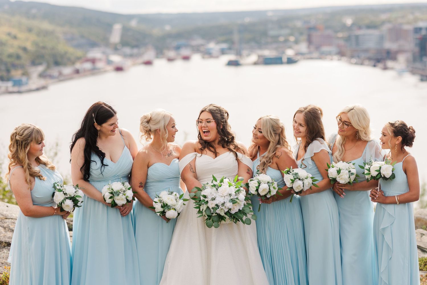 Row of people wearing light blue bridesmaid dresses stand together holding white bouquets at an outdoor wedding event.