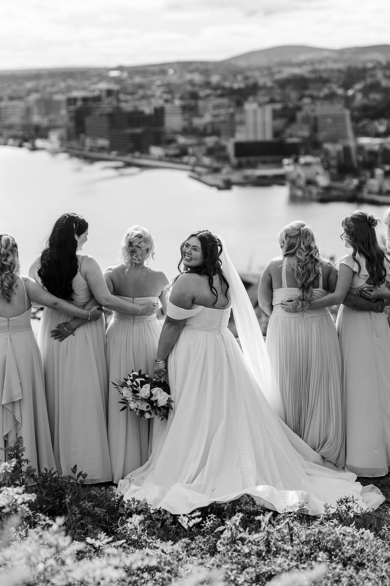 A wedding party wearing light-colored gowns stands in a row against a city backdrop, viewed from behind in black and white.