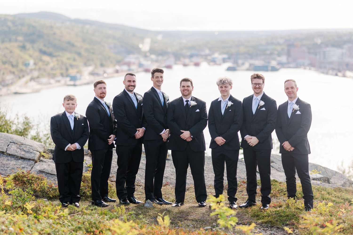Groomsmen in black suits pose together on a scenic hill overlooking the mountains during a wedding photoshoot.