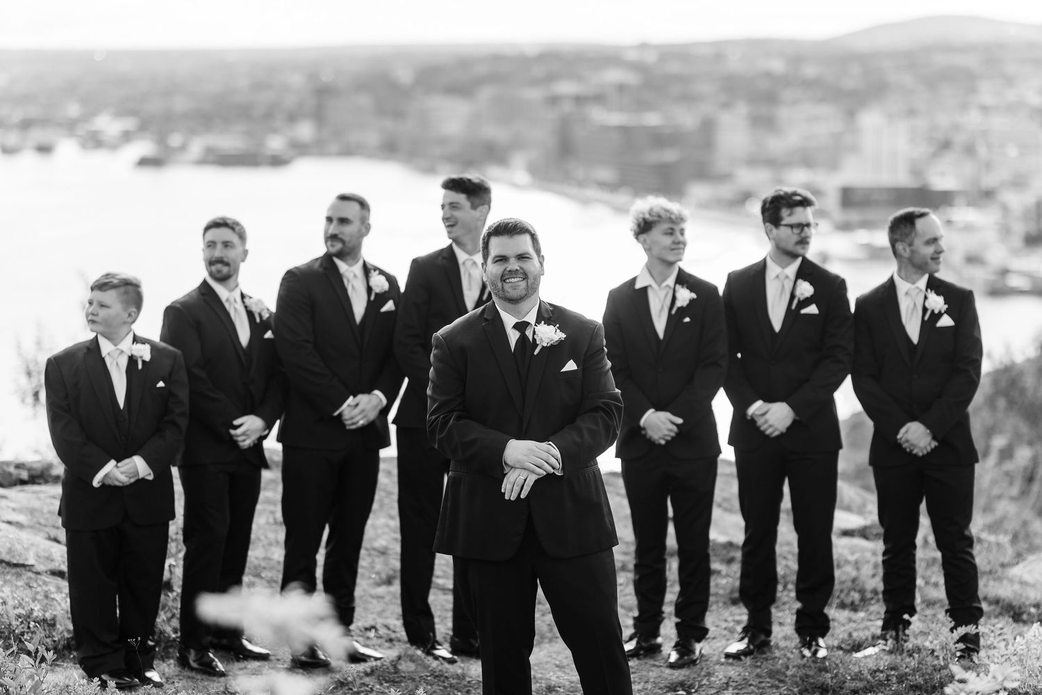A wedding party in black suits stands on a hilltop overlooking a scenic mountain landscape in black and white photography.