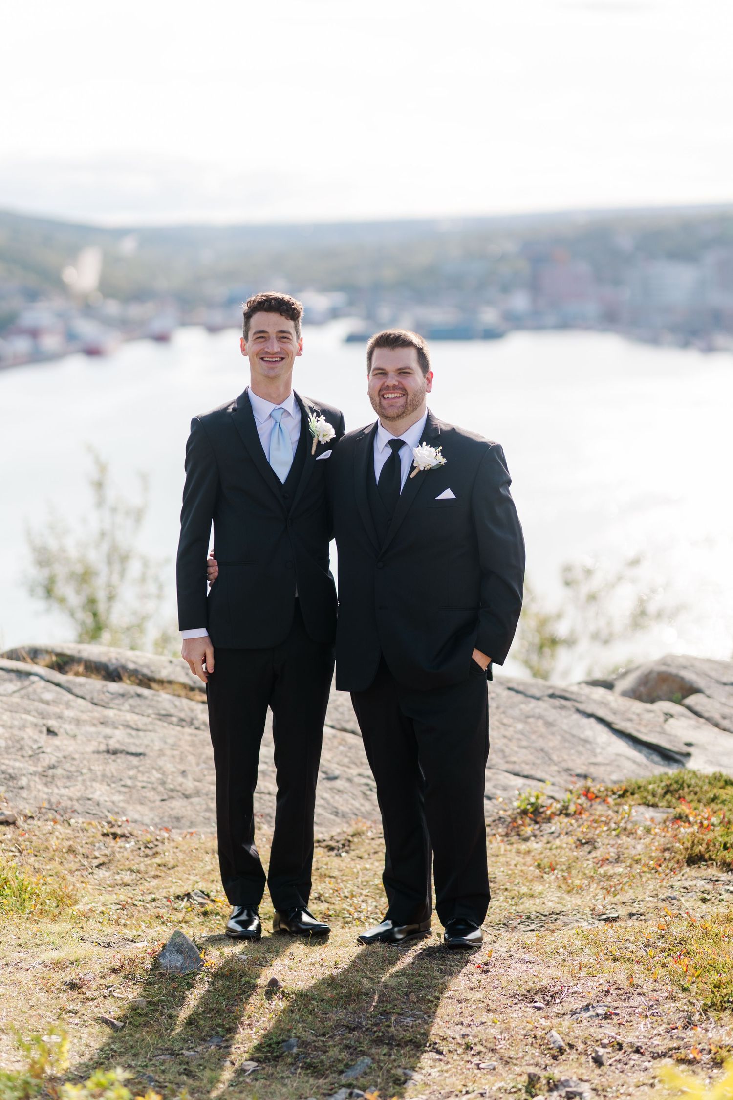 Two men in black tuxedos stand together on a rocky cliff overlooking a scenic coastal landscape.