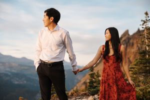 Couple in formal attire poses romantically against mountain backdrop with woman in flowing red dress.