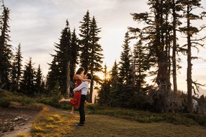 A person stands admiring the golden sunset through tall evergreen trees in a mountainous landscape.