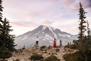 Group of hikers stand along a rocky mountain ridge with Mount Rainier in the background during sunset.