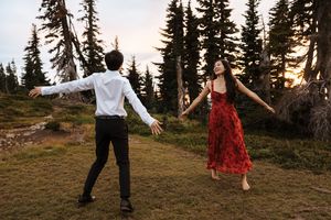 Two people in formal attire dance joyfully in a mountain forest setting during golden hour sunset.