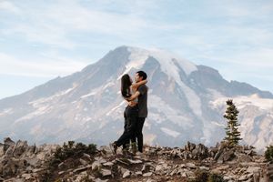 A couple embraces on a rocky overlook with snow-capped Mount Rainier rising majestically in the background.
