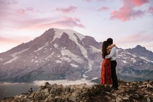 A dramatic pink sunset illuminates snow-capped mountain peaks and a hiker silhouetted against the alpine landscape.