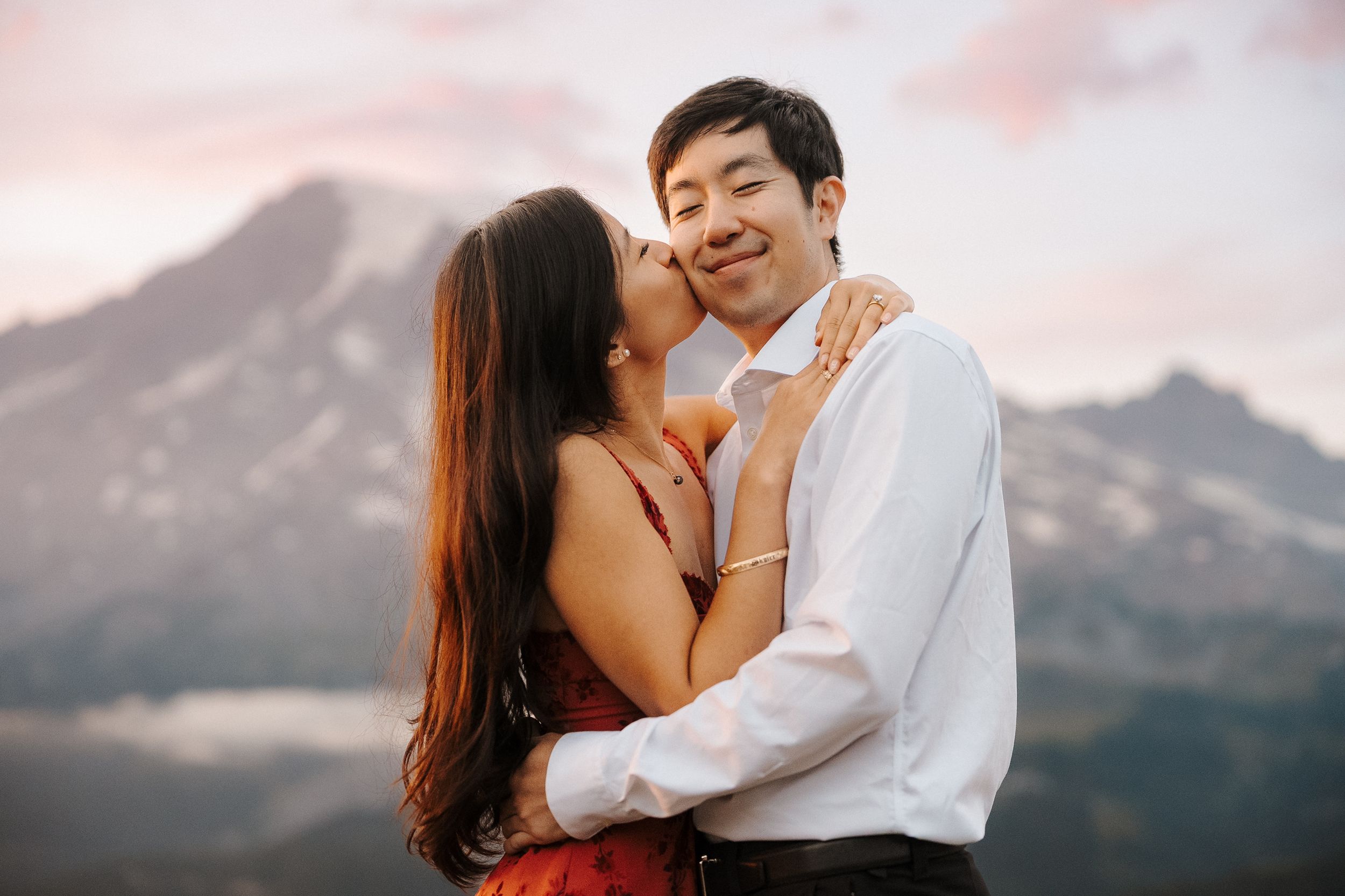 A couple in white embraces romantically against a mountain backdrop during sunset.